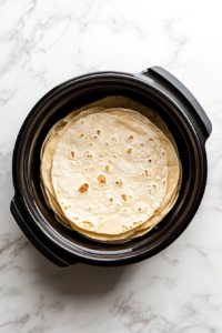 This image shows tortillas being placed in the bottom of a slow cooker, forming the first layer for the Crockpot Enchiladas.