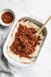 This image showcases layers of vegan lasagna sheets being topped with rich plant-based bolognese and creamy béchamel sauce in a baking dish.