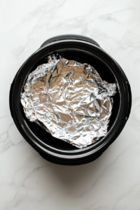 This image shows the interior of a slow cooker being carefully lined with aluminum foil, ensuring easy cleanup and even cooking for the meatloaf.