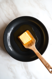 top-down view of a black skillet on a white marble countertop with a stick of unsalted butter melting into a glossy golden liquid. A wooden spatula rests beside the skillet, ready for stirring.