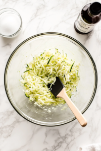 top-down shot of grated zucchini being mixed into a bowl of wet ingredients, including eggs and oil, for the zucchini bread batter.