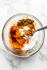 A bowl filled with Greek yogurt, lime juice, chili powder, turmeric, and other spices being mixed together to create a vibrant tandoor marinade.