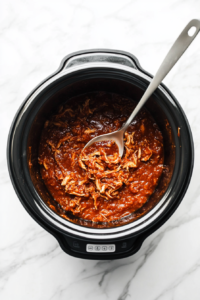 Shredded chicken being mixed back into the slow cooker with the tangy BBQ sauce, ensuring every piece is coated for maximum flavor.