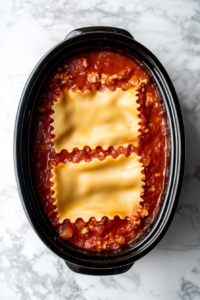 This image shows uncooked lasagna noodles being carefully placed over a layer of marinara sauce in the slow cooker to start building the lasagna.