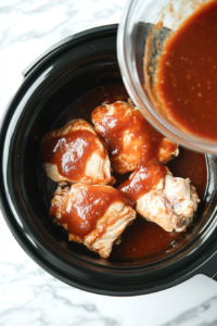 The tangy and flavorful BBQ sauce being poured over the chicken breasts in the slow cooker, ensuring the chicken is coated evenly for slow cooking.