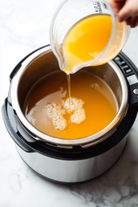 measuring cup filled with chicken broth being poured into the bottom of an Instant Pot, creating a flavorful base for cooking the seasoned keto chicken.