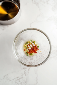 This image shows hot oil being poured over a bowl of minced garlic and chopped spring onions, sizzling and releasing a burst of aromatic flavors to form the garlic noodle sauce base.