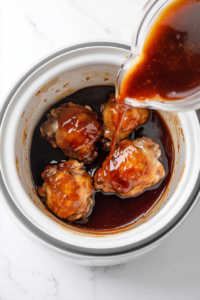 This image shows a thick, savory sauce of soy sauce, ketchup, honey, garlic, and basil being poured over raw chicken thighs placed inside a slow cooker for cooking.