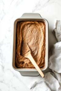 This image shows the second half of the bread batter being poured carefully over the cinnamon-sugar layer in the loaf pan, forming the top layer of the cinnamon swirl bread.