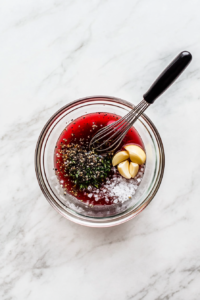 This image shows the ingredients for the salad dressing being whisked together in a separate bowl, blending olive oil, red wine vinegar, lemon juice, garlic, oregano, salt, and black pepper.