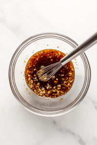 This image shows a bowl with orange sauce ingredients being whisked together, including orange juice, soy sauce, garlic, and other seasonings.