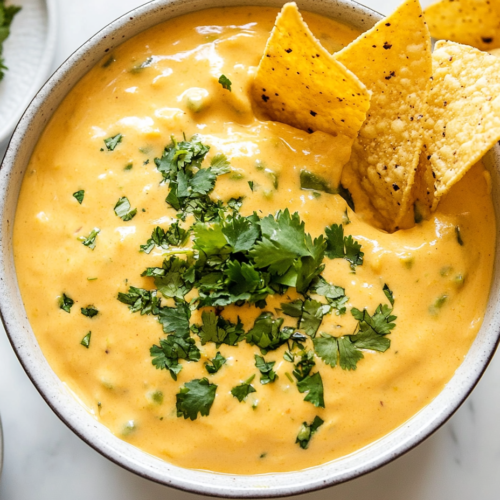 this image shows A creamy queso dip served in a white bowl, topped with freshly chopped cilantro and seasoned with hints of cumin, red pepper flakes, and chili powder for a rich, flavorful appetizer.