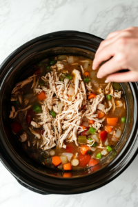 This image shows shredded chicken being added back to the slow cooker, mixing into the broth and vegetables.