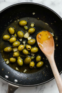 this image shows A pan showing green olives and minced garlic being sautéed, releasing aromatic flavors for the Vegan Green Olive Pasta Sauce.