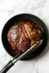 This image shows a seasoned chuck roast being seared in a hot skillet, creating a golden-brown crust to enhance the flavor of the pot roast.