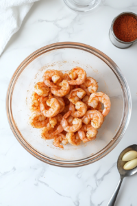 This image shows shrimp in a bowl being seasoned with paprika, minced garlic, and pepper, ensuring each piece is evenly coated for the flavorful dish.