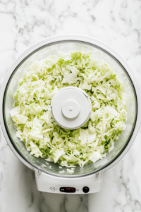 This image shows cabbage being shredded in a food processor, turning it into finely shredded pieces that will be used as a low-carb rice substitute.