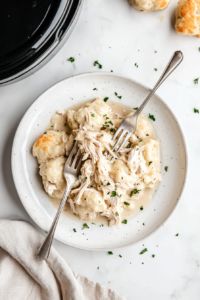 This image shows tender, slow-cooked chicken being shredded with two forks, making it ready to be stirred back into the creamy dumplings mixture.