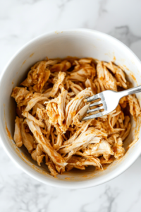 This image shows cooked chicken being shredded with two forks on a cutting board, creating tender pieces for the Crockpot Chicken Noodle Soup.