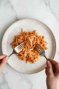 This image shows cooked chicken being shredded using two forks, creating tender, flavorful pieces to be added back into the Crockpot White Chicken Chili.