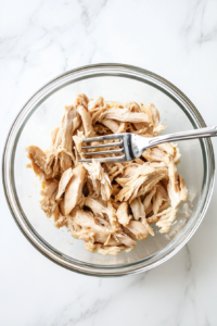 This image shows a pair of forks shredding the tender cooked chicken into small pieces, ready to be mixed back into the creamy pot pie filling.