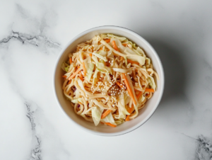 This image shows a white bowl filled with simple oriental Asian cabbage salad, made with shredded cabbage, carrots, toasted sesame seeds, and crunchy ramen noodles, tossed in a flavorful dressing.