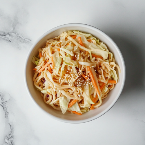 This image shows a white bowl filled with simple oriental Asian cabbage salad, made with shredded cabbage, carrots, toasted sesame seeds, and crunchy ramen noodles, tossed in a flavorful dressing.