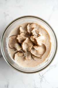 this image shows Oyster mushrooms soaking in a creamy vegan buttermilk mixture to absorb flavor and prepare for the flour coating.