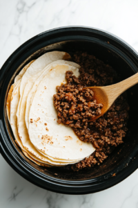 This image shows the seasoned ground beef mixture being evenly spread over the tortillas in the slow cooker as part of the Crockpot Enchiladas layering process.