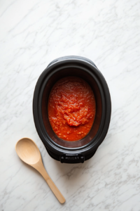 This image shows marinara sauce being evenly spread in the bottom of a slow cooker, creating the flavorful base for the crockpot lasagna layers.