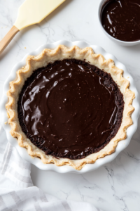 This image shows a thick layer of chocolate brownie batter being evenly spread over the golden-brown pie crust, ready for its final bake.