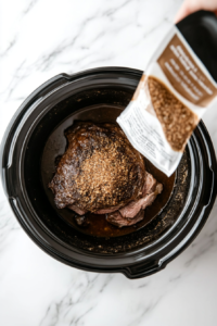 This image shows onion soup mix being evenly sprinkled over the seared chuck roast in the slow cooker, adding depth and savory flavor to the dish.