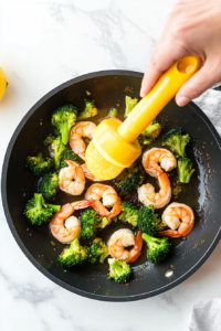 This image shows fresh lemon juice being squeezed over shrimp and broccoli in the skillet, adding a zesty and tangy flavor to the dish.