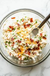 This image shows the chicken salad being stirred with a large spoon to evenly distribute the bacon and cheese throughout the creamy mixture.