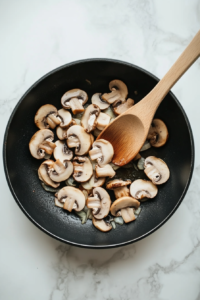 this image shows Sliced mushrooms being stirred into a skillet with sautéed onions and garlic, starting to brown and release their earthy flavors for the stroganoff.