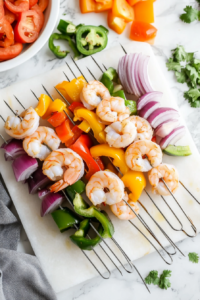 This image shows raw jumbo shrimp, onion slices, and bell pepper pieces being threaded onto long wooden skewers, ready for grilling.