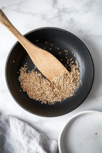 This image shows sesame seeds being toasted in a small skillet over medium heat until golden and fragrant, an essential step for enhancing the salad's flavor.