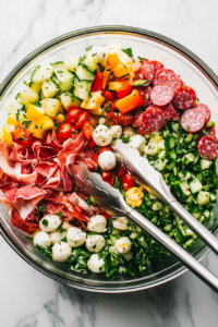 This image shows a large spoon and fork tossing the meats, cheese, vegetables, and dressing together in a salad bowl, ensuring all ingredients are evenly coated.