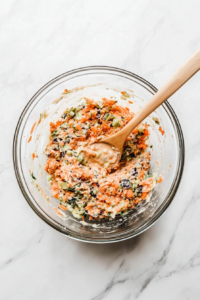 this image shows bowl of shredded cabbage, carrots, and green onions being tossed with a creamy, tangy keto dressing, evenly coating the vegetables for a flavorful coleslaw.