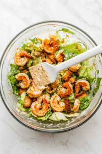 This image shows the ingredients for the shrimp Caesar salad—shrimp, chopped romaine, croutons, and dressing—being tossed together in a large bowl.