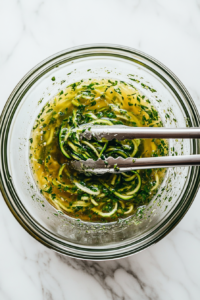 This image shows zucchini noodles being tossed with a freshly made Greek dressing in a large bowl, coating each strand for a flavorful base.