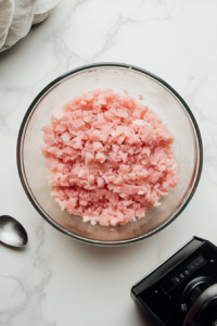 This image shows finely chopped ham being transferred from a food processor to a mixing bowl, ready for combining with other ingredients.