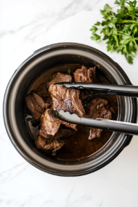 This image shows the perfectly seared chuck roast being lifted from a skillet and transferred into the slow cooker to begin the pot roast preparation.