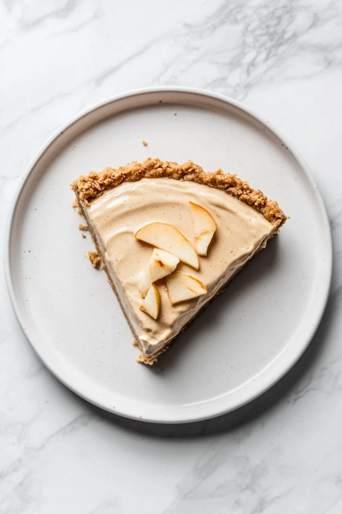 this image shows delicious vegan apple cream pie with a golden-brown crust, creamy filling, and fresh apple slices elegantly arranged on top, served on a white round plate.