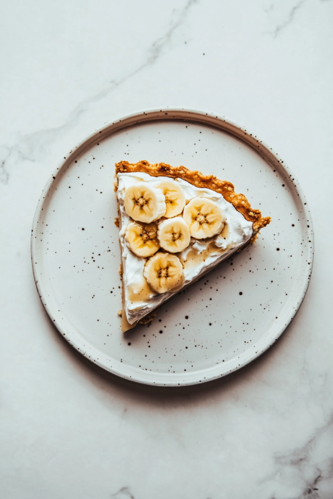 this image shows luscious slice of vegan banana cream pie on a white plate, topped with fresh banana slices and a creamy layer of banana-flavored filling.