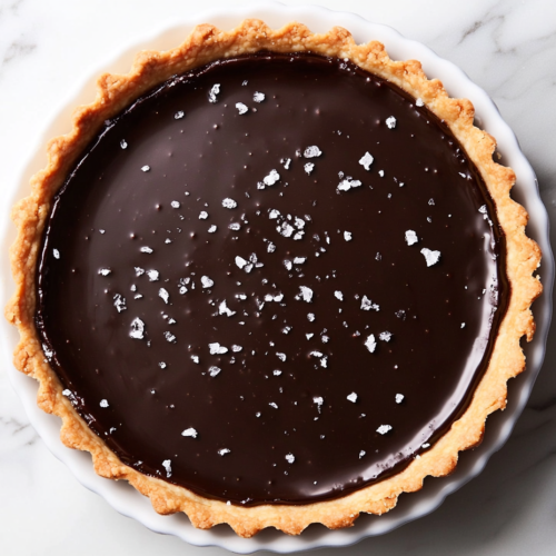 This image shows a rich and glossy vegan dark chocolate tart in a round dish, with the smooth chocolate filling perfectly set in a golden crust, ready to serve as an indulgent dessert.