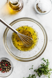 This image shows a small bowl with olive oil, white wine vinegar, sea salt, and black pepper being whisked together to create a tangy and flavorful dressing for the antipasto salad.