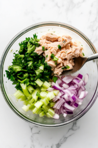 This image shows a top-down view of additional ingredients such as diced vegetables, herbs, or seasonings being added to the tuna and mayonnaise mixture to enhance flavor and texture.