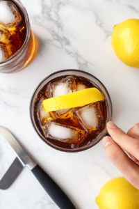 This image shows a lemon twist being placed on the rim of the glass, enhancing the Whiskey and Coke with a touch of citrus aroma and flavor.