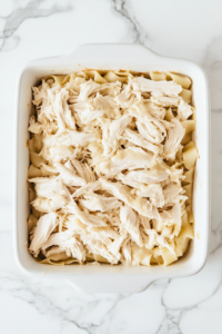 This image shows shredded buffalo chicken being evenly spread over the pasta layer, adding the signature spicy flavor to Buffalo Chicken Lasagna.